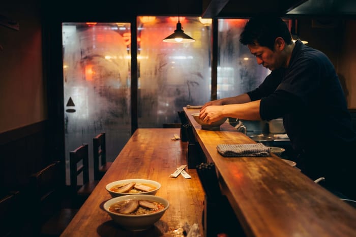 A chef working at the pass of a fourteen-seat ramen counter at night, steam on the window behind