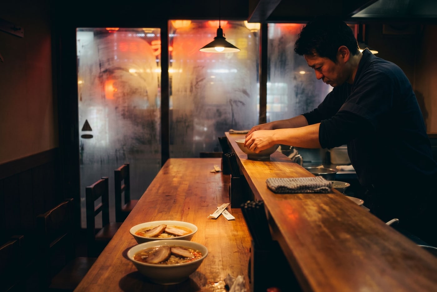 A chef working at the pass of a fourteen-seat ramen counter at night, steam on the window behind